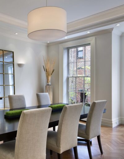 Elegant dining room with a large wooden table, upholstered chairs, and a view of exterior greenery through the windows.