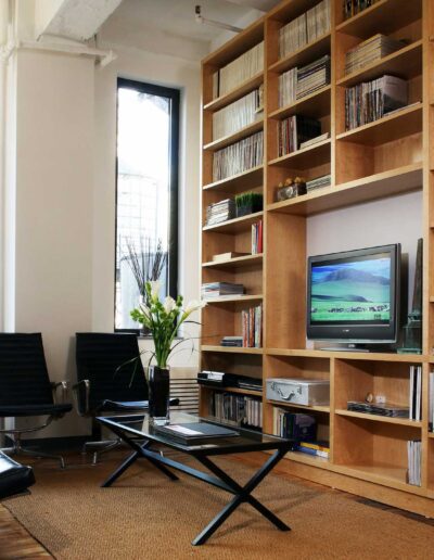 A modern living room with a large bookshelf, flat screen tv, and two black leather chairs, featuring tall windows with a view of urban buildings.