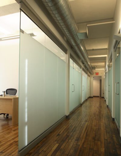 An empty office corridor with glass partition walls and wooden flooring.