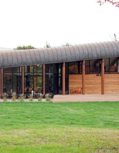 Modern wooden building with curved metal roof surrounded by greenery.