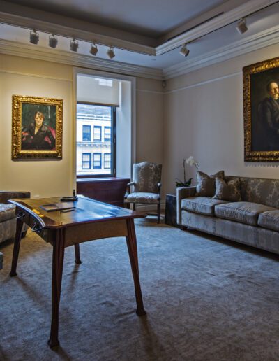 An elegant room with classic paintings and antique-styled furniture overlooking a city street through large windows.