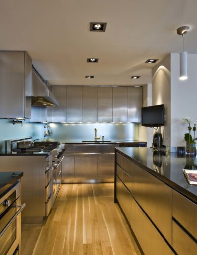 Modern kitchen interior with stainless steel appliances, polished wooden floors, and a neatly arranged fruit bowl on the counter.